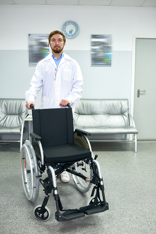 Portrait of young bearded doctor standing in hospital hall posing with empty wheelchair and