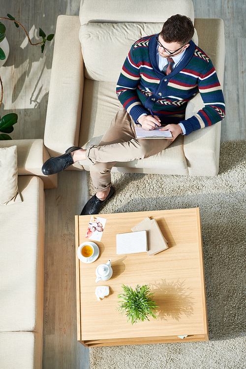 Above view portrait of pensive male psychologist writing notes on clipboard while sitting in comfortable armchair in sunlit office, copy space