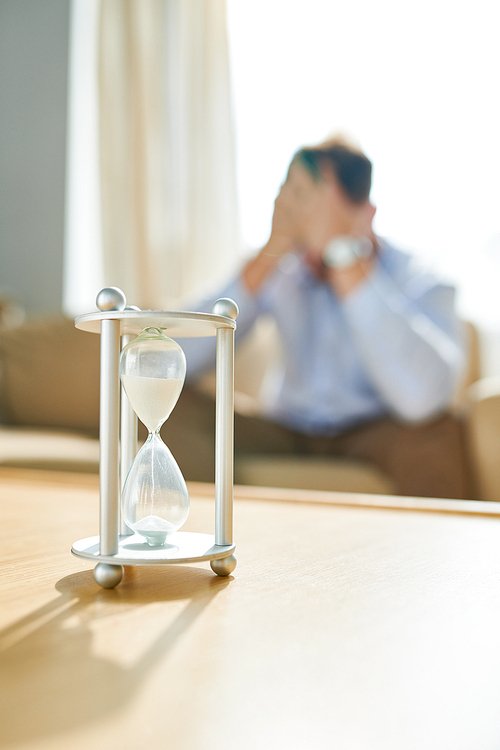 Closeup of hourglass on table with blurred shape of stressed man waiting in background, copy space