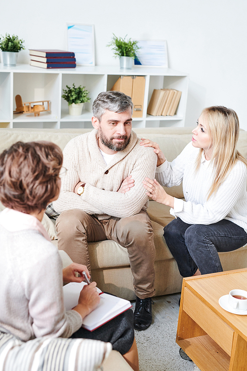 Serious pensive middle-aged husband with gray hair sitting on sofa and being distant while listening to pretty wife at therapy session, young woman touching his shoulder and supporting him