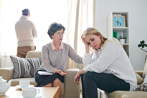 Understanding mature lady psychologist in glasses stroking shoulder of young woman while supporting her at therapy session while her husband looking out window in background, relationship difficulties concept