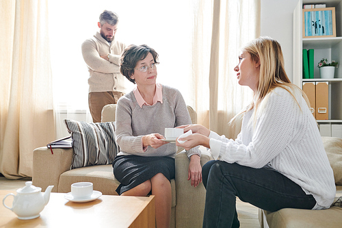 Careful mature psychologist in sweater sitting in armchair and giving cup of tea to patient while supporting young woman having problems in marriage at therapy session for couple.