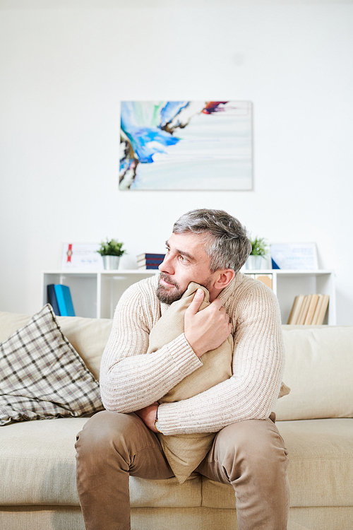 Emotionally instable middle-aged bearded man with gray hair sitting on sofa and embracing cushion while talking to psychologist in modern office room