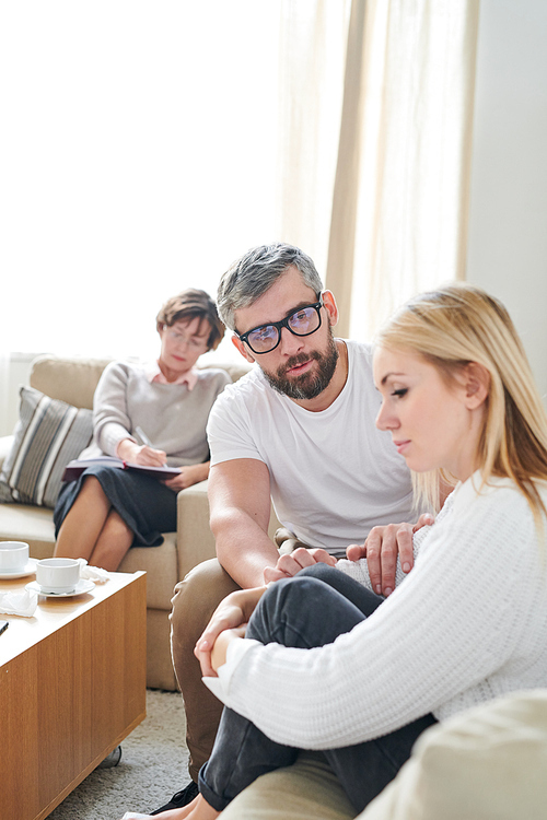 Sad young wife feeling sorrow sitting on sofa and embracing her legs while her husband consoling her at therapy session