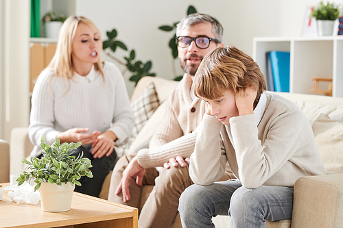 Sad depressed teenage son in sweater sitting on sofa and covering ears to avoid parents quarrel while his parents fighting in background in living room