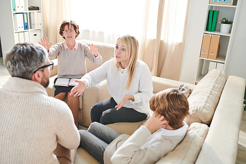 Aggressive parents sitting on sofa and fighting in front of teenage son while mature psychologist trying to stop conflict at therapy session