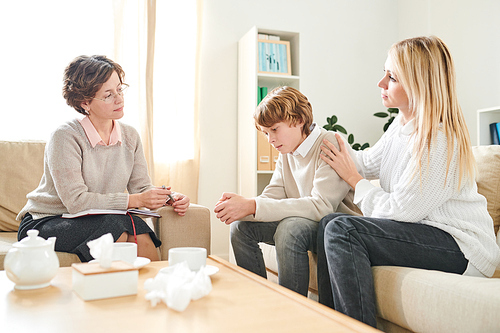 Serious mother worrying about teenage son and holding his shoulder while supporting him at therapy session with psychologist, depressed boy coping with his complexes