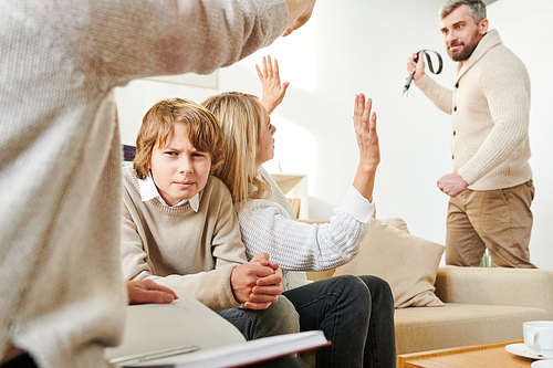 Sad frowning boy suffering from violence in family: angry bearded man holding belt while biting wife in front of son and psychologist