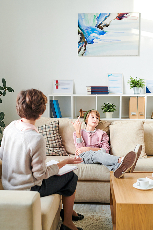 Serious indifferent teenager in hat sitting on sofa and keeping feet on table while fucking off psychiatrist at therapy session