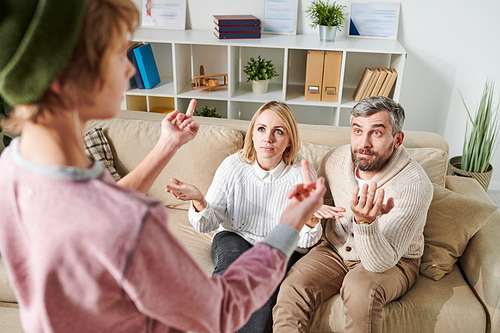 Troubled teenager in hipster clothing standing in front of parents and showing middle finger while fucking off them, shocked parents shrugging arms and expressing his displeasure