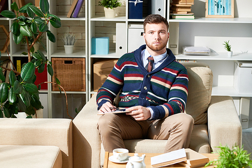 Portrait of handsome  psychologist posing sitting in chair and  in therapy office, copy space