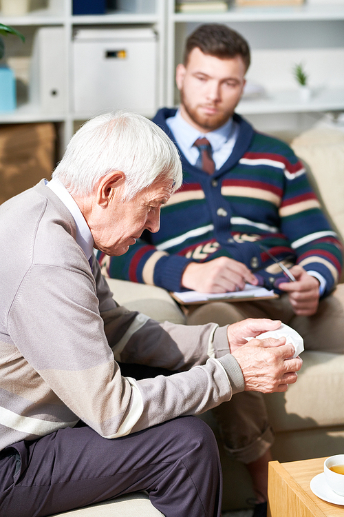 Serious pensive senior man sitting on sofa and looking down while telling about his fears with shrink, he holding napkin to wipe tears