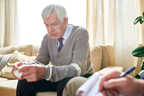 Serious handsome elderly man with wrinkles wearing cardigan sitting on sofa and holding napkin while talking to psychiatrist about his problems at therapy