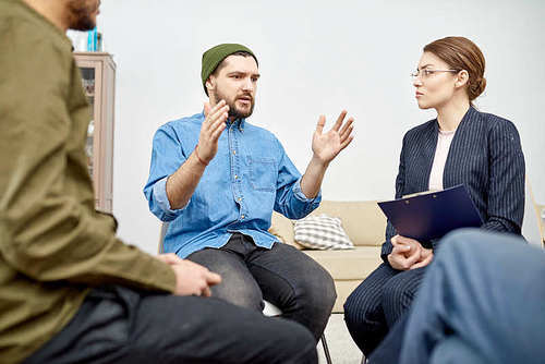 Bearded patient wearing denim shirt expressing his feelings while sitting in circle and participating in group therapy session, pretty psychiatrist listening to him with concentration