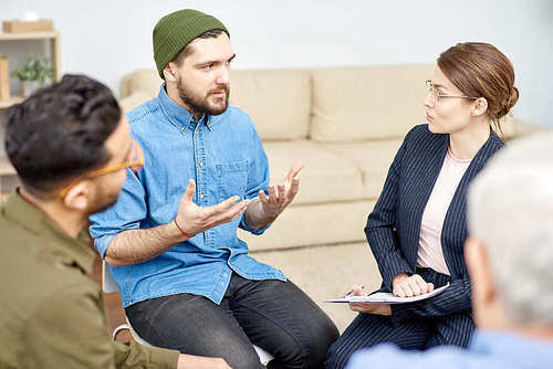 Highly professional young psychologist listening to her male patient with concentration while guiding support group suffering from phobias, interior of cozy office on background