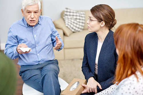 Senior man sitting in circle of psychological support group and sharing his problems with mentor, other patients listening to him with concentration