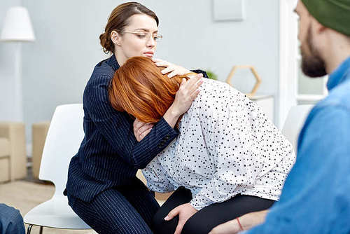 Beautiful young psychiatrist comforting her crying obese patient suffering from eating disorder while conducting group therapy session at cozy office