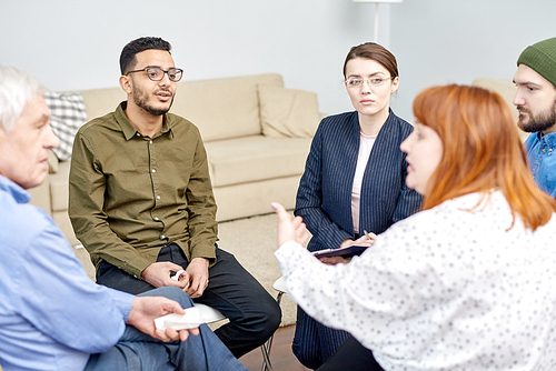 Profile view of red-haired obese woman discussing faced problem with other patients while participating in group therapy session, pretty psychologist listening to her with concentration