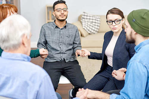 Multi-ethnic group of patients suffering from phobias sitting in circle and holding hands while highly professional psychologist conducting session, interior of cozy office on background