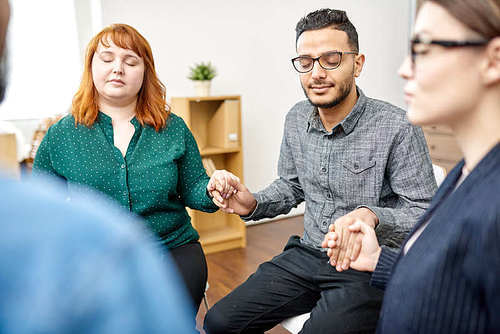 Male and female patients with closed eyes sitting in circle and holding hands while participating in group therapy session, interior of cozy psychotherapy office on background