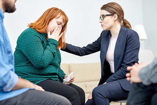 Highly professional psychiatrist comforting her crying obese patient suffering from eating disorder while conducting group therapy session at cozy office