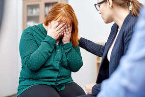 Friendly young psychiatrist in eyeglasses supporting crying woman suffering from eating disorder while conducting group therapy session at cozy office