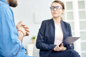 Highly professional young psychiatrist with clipboard in hands sitting in circle while listening to member of rehab group at therapy session