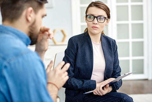 Depressed bearded man having face to face problem discussion with talented psychologist while sitting at cozy office, she holding clipboard in hands and listening to him with concentration