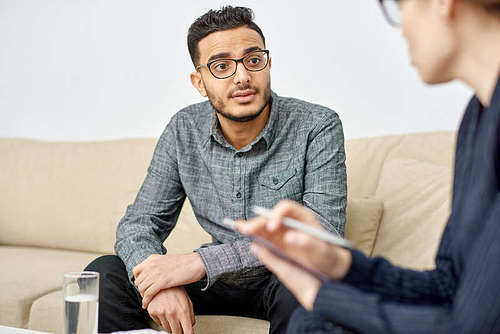 Handsome young man suffering from depression sitting on cozy sofa and listening to highly professional psychologist while she giving him piece of advice
