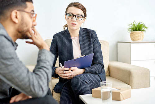 Profile view of handsome young man sitting on sofa while having consultation with highly professional psychiatrist at cozy office