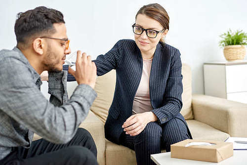 Profile view of sad mixed-race man drinking water while trying to cope with grief, friendly young psychologist encouraging him while having appointment at modern office