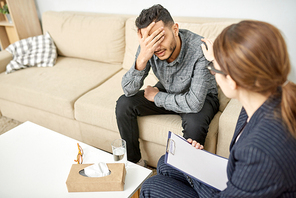 Despaired young patient covering face with hand while trying to cope with grief during therapy session at modern office, friendly psychiatrist encouraging him