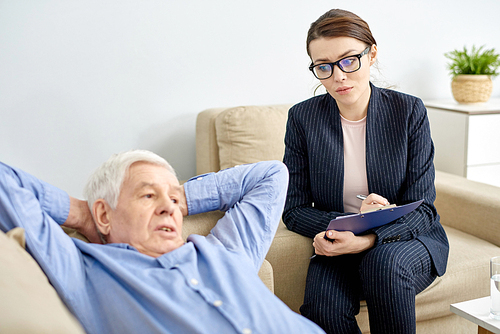 Depressed senior man lying on cozy sofa of psychotherapy office and talking to highly professional young psychologist, she taking necessary notes