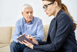 Profile view of attractive young psychologist taking necessary notes while having appointment at modern office, senior patient