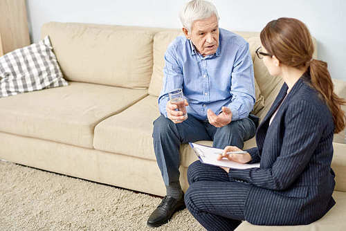 Senior patient with glass of water in hand sitting on cozy sofa and discussing faced issue with highly professional psychologist while having therapy session