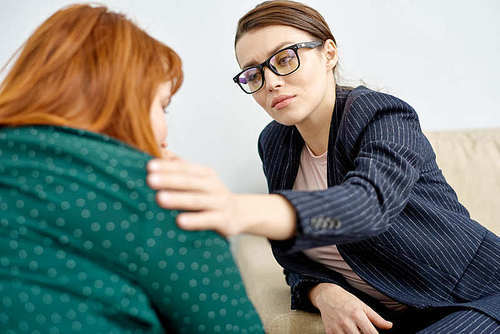 Friendly young psychiatrist wearing suit and eyeglasses comforting crying obese patient while conducting therapy session
