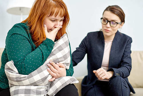 Highly professional psychiatrist wearing elegant suit comforting crying obese patient while conducting therapy session, office interior on background