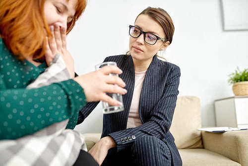 Crying obese woman with glass of water in hand expressing her feelings while sitting on cozy couch of psychotherapy office, friendly specialist comforting her