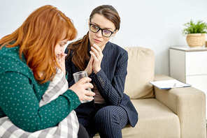 Profile view of crying obese woman holding glass of water in hand while sharing problems with talented young psychologist, interior of cozy office on background