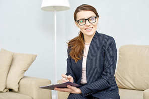 Beautiful young psychologist wearing suit and eyeglasses  with charming smile while taking notes on clipboard, interior of cozy office on background