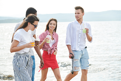 Group of travelers walking along the seaside drinking cocktails and discussing their vacation