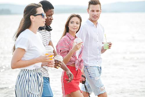 Group of friends with fresh juice discussing while walking along the seaside outdoors
