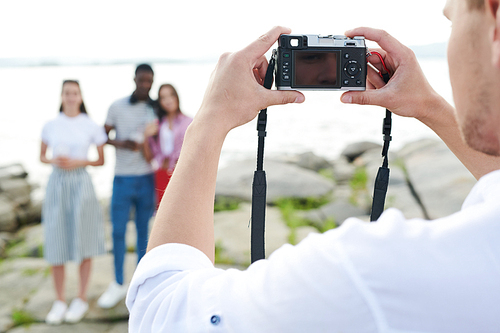 Rear view of young man holding digital camera and photographing young people on the seaside