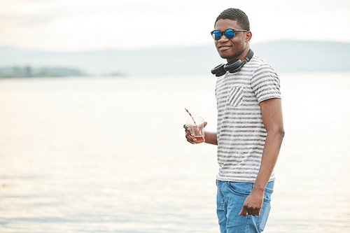 Portrait of smiling African man wearing sunglasses standing in front of the sea and drinking cocktail