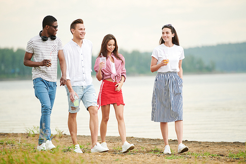 Group of stylish young people with drinks walking along the lake and communicating with each other
