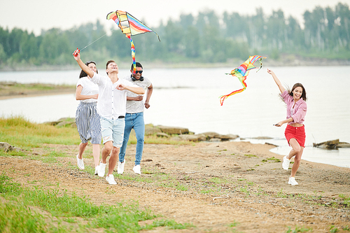 Group of young people running along the lake with colorful flying kite
