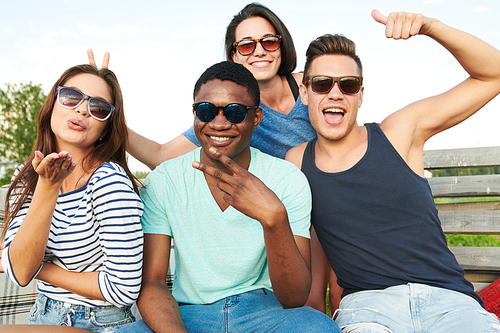 Group of happy young people in sunglasses sitting on bench and posing to the camera