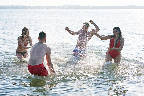 Cheerful hilarious young friends in swimwear laughing while splashing water enjoying summer vacation at sea