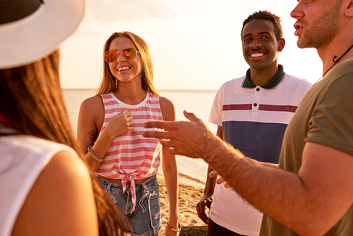 Smiling young multiethnic friends in casual clothing discussing summer vacation while gathering together on beach: pretty girl in sunglasses and African guy attentively listening to friends story