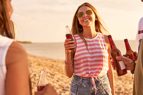 Cheerful optimistic beautiful young woman in sunglasses holding bottle of soda smiling happily while talking to friends at beach party